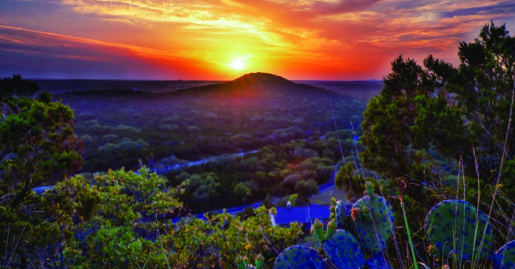 Vibrant sunset casting an orange glow over the lush, rolling hills and valleys of the Texas Hill Country near Wimberley, TX, with prickly pear cactus plants in the foreground.