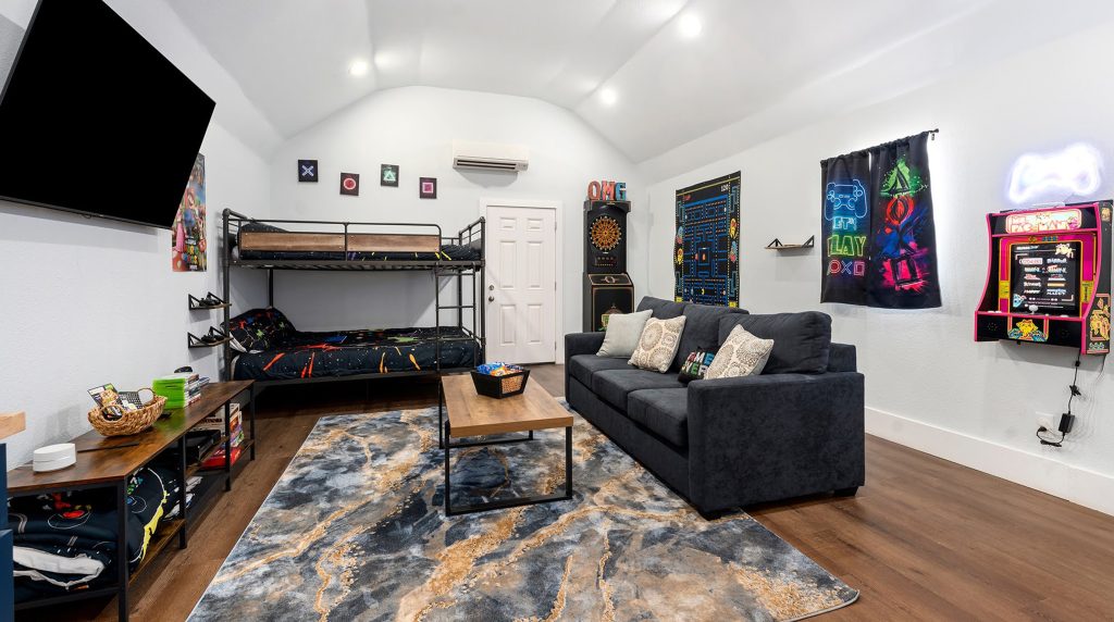 A detailed view of a modern game room within a Wimberley, TX vacation rental, featuring a sofa, coffee table, bunk beds, arcades, and a large TV, illustrating cozy and fun romantic things to do in Wimberley, TX.