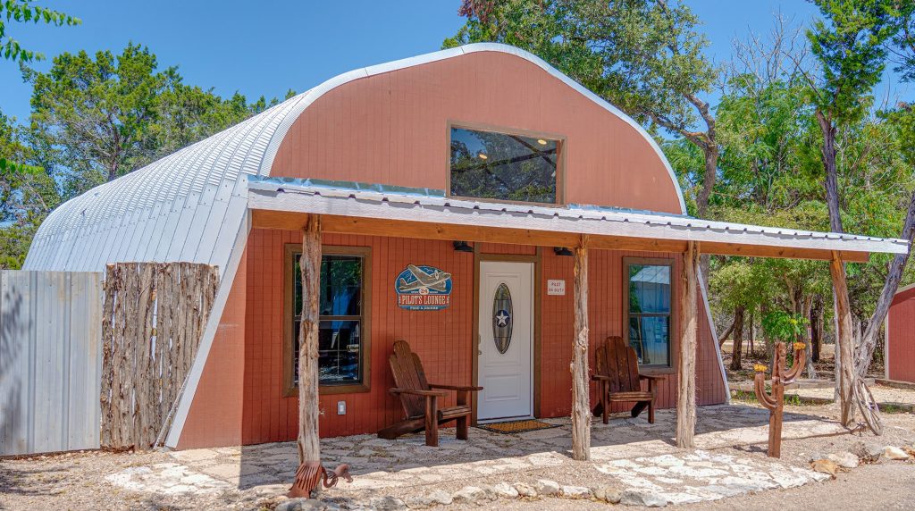 A charming red aviation-themed cabin rental with a curved metal roof and rustic wooden porch chairs, perfect for couples looking for romantic things to do in Wimberley, TX.