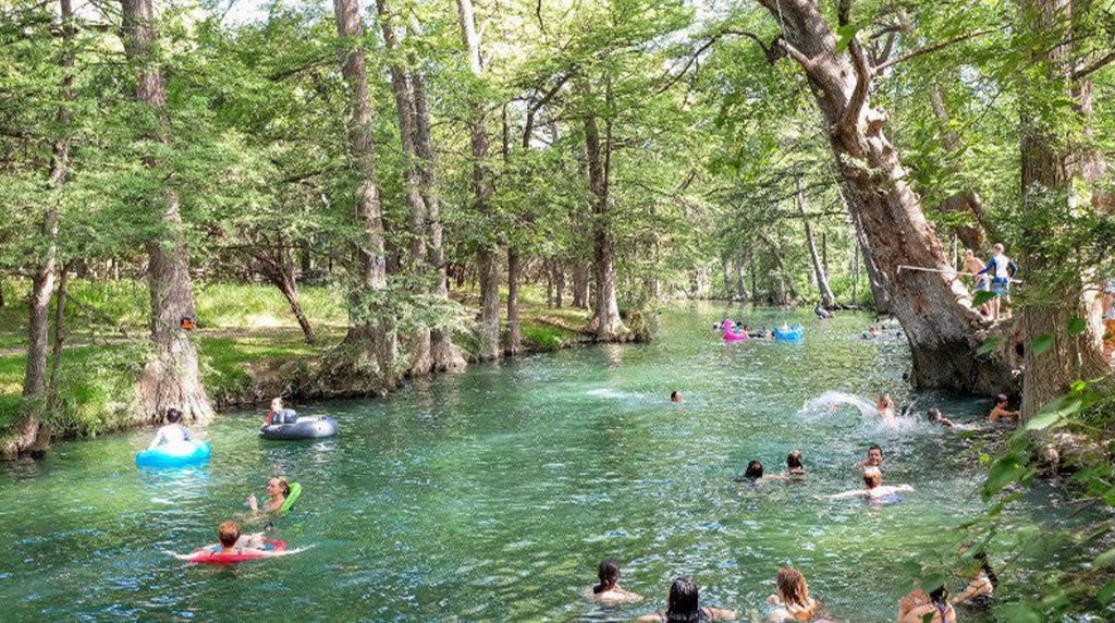 Swimmers and tubers enjoying Cypress Falls Swimming Hole under tall trees.